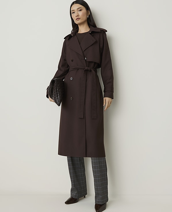 A woman in a brown trench coat and plaid pants stands against a plain white background, holding a dark clutch.