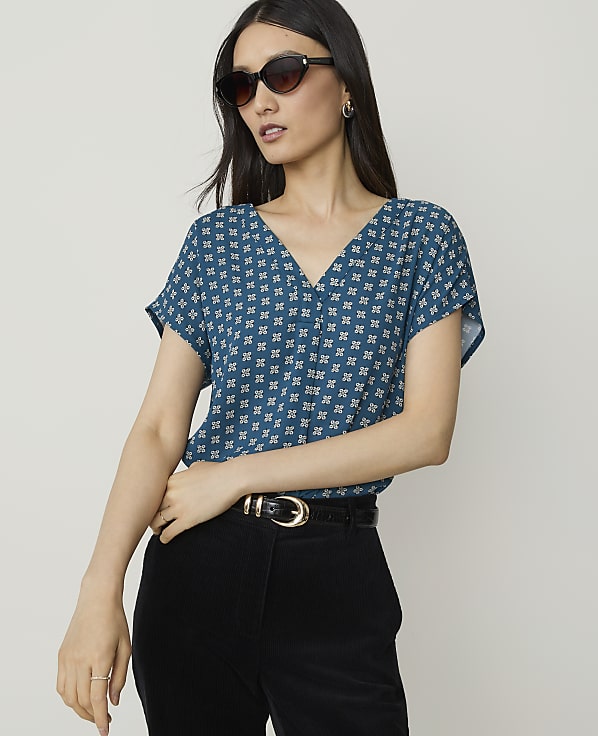 A woman in a patterned teal top and black pants stands against a plain, light background.
