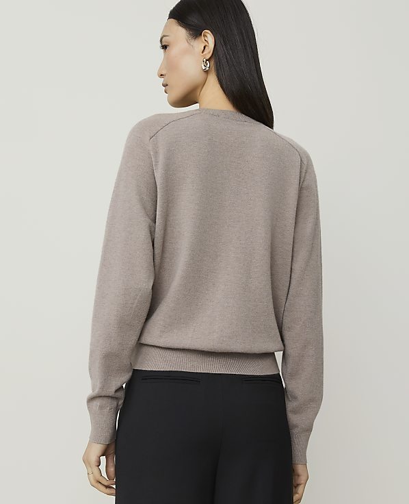 A woman in a taupe sweater and black pants stands against a plain, light gray background.