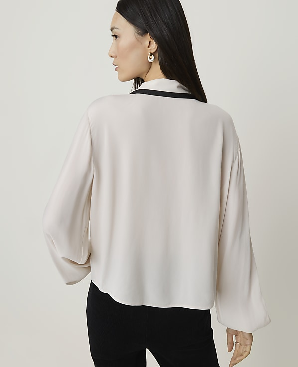 A woman in a cream blouse and black pants stands against a plain, light background.