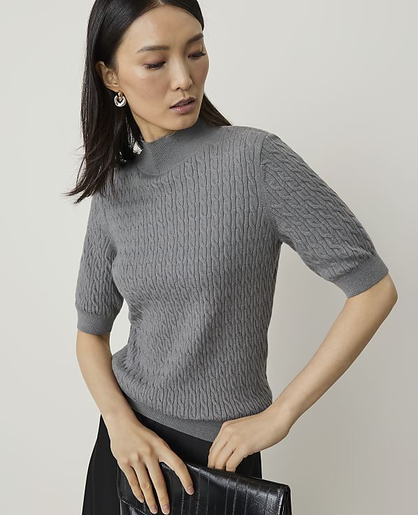 A woman in a grey cable-knit sweater and black skirt holds a black clutch in front of a plain, light-colored background.