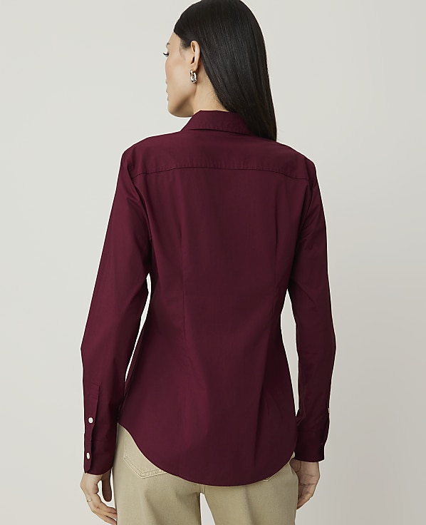 A woman in a maroon button-down shirt and tan pants stands against a plain, light gray background.