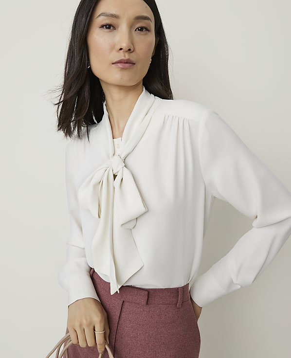A woman in a white pussy-bow blouse and pink trousers stands against a plain, light background.