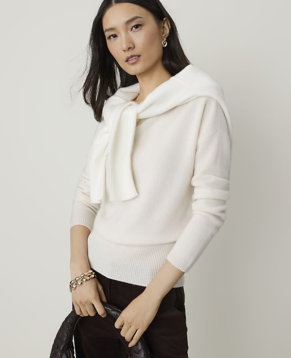 A woman in a cream sweater and dark pants holds a woven bag in front of a plain background.