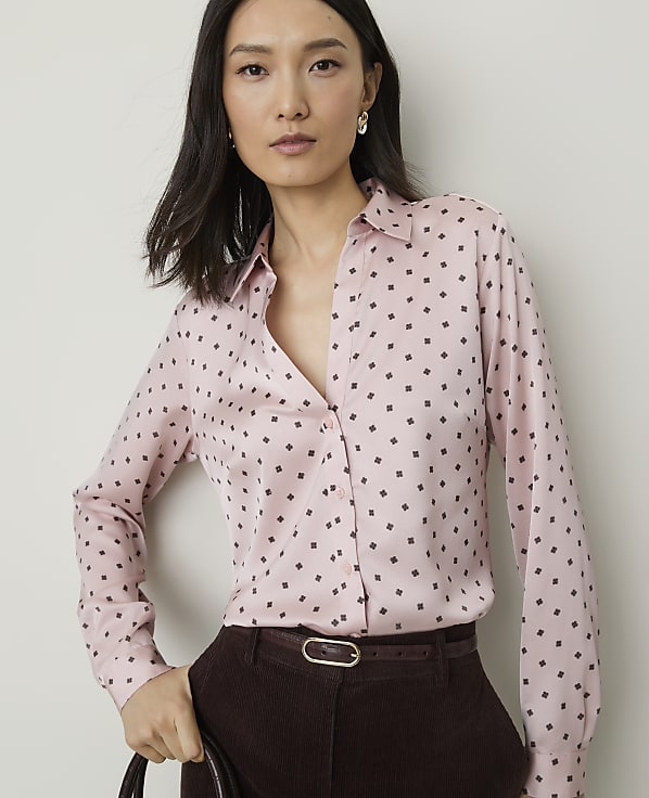 A woman in a pink patterned blouse and brown pants stands against a plain white background.