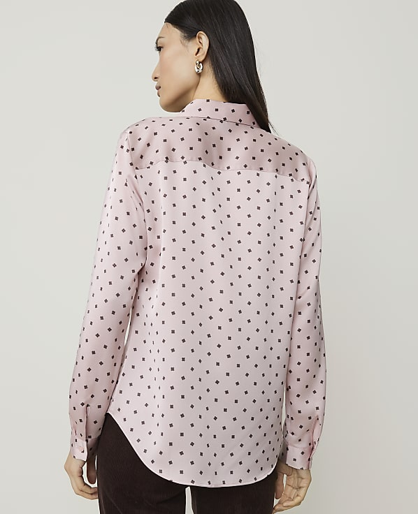 A woman in a pink, polka-dotted blouse stands against a plain, light-colored background.