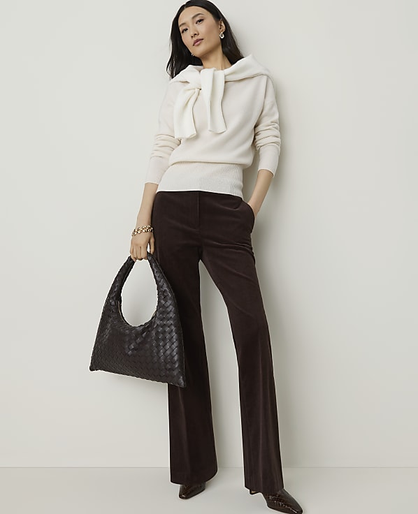A woman in a cream sweater and brown corduroy pants stands with a dark woven bag against a plain white background.