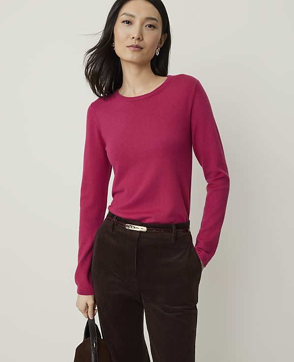 A woman in a pink sweater and brown corduroy pants stands against a plain white background, holding a dark bag.