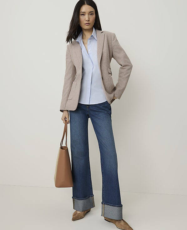 A woman in a light brown blazer, blue shirt, and cuffed jeans stands against a plain, light background, holding a tan tote bag.