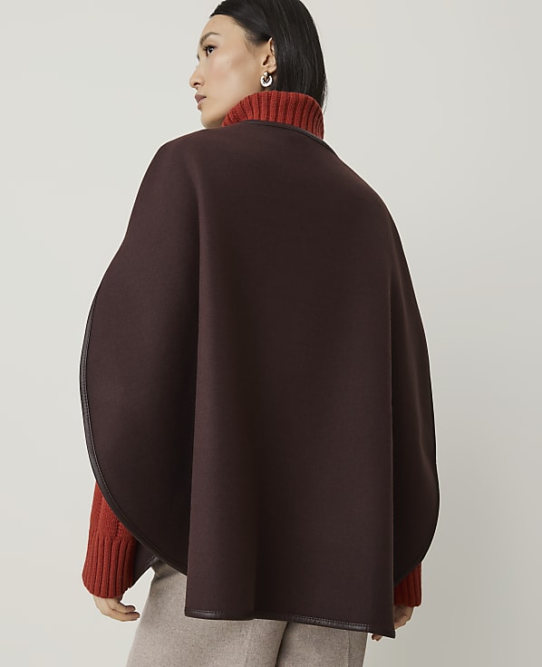 A woman in a brown cape and orange turtleneck stands against a plain, light-colored background.