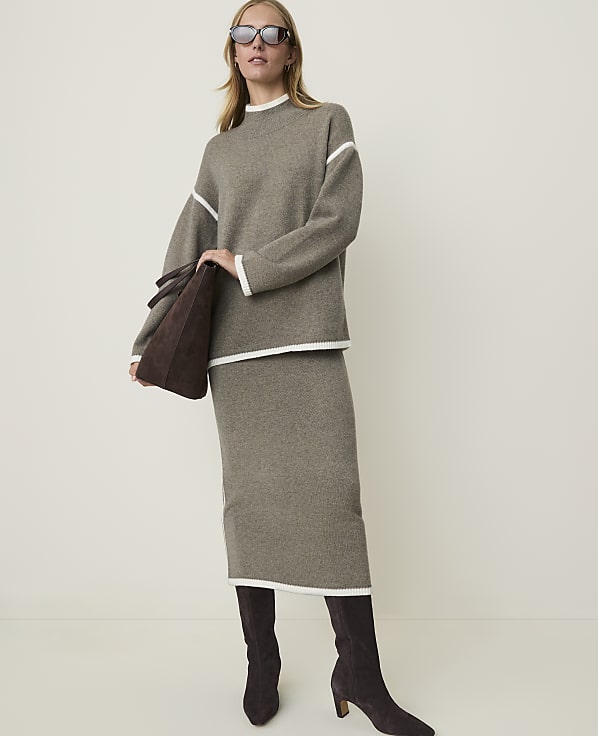 A woman in a taupe knit sweater and skirt with white trim, holding a brown suede tote bag, stands against a plain, light-colored background.