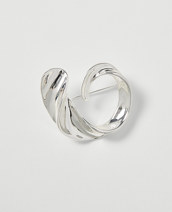 A silver, abstractly shaped brooch with a pin is centered on a plain white background.