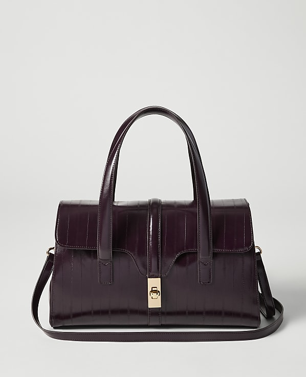 A deep purple, croc-embossed handbag with a gold clasp and top handles sits in the foreground against a plain, light gray background.