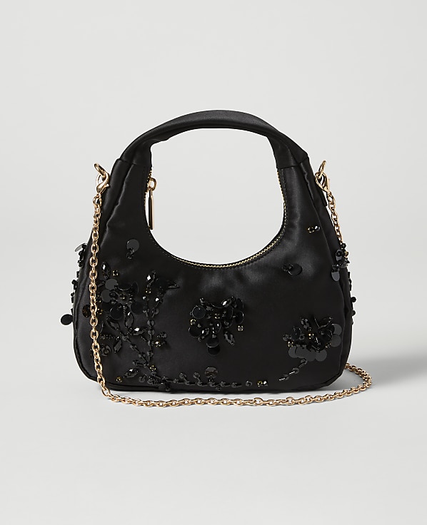 A black satin handbag adorned with black sequins and beads sits on a light gray background, with a delicate gold chain strap draped across it.