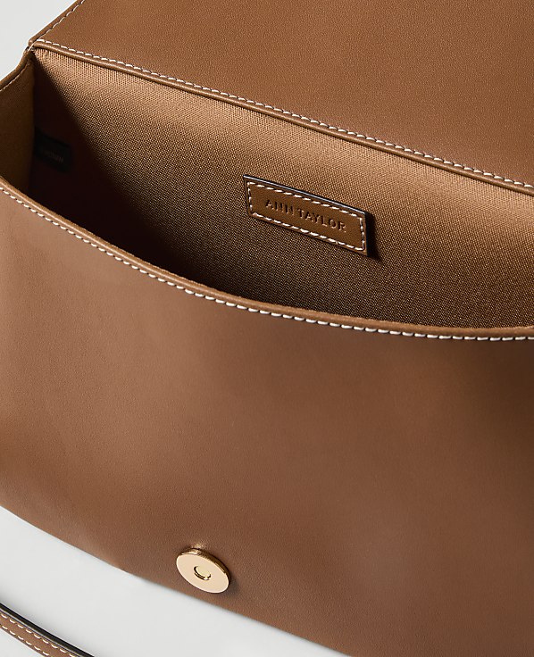 A brown leather clutch with a gold clasp is open, revealing its satin interior and a brand tag, against a plain white background.