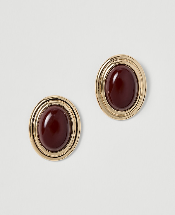 A pair of oval, gold-toned earrings with deep red cabochon stones are displayed against a plain, light gray background.