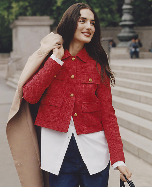 A woman in a red jacket and white shirt walks in front of a blurred background of stone steps and people.