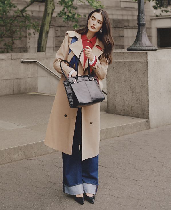 A woman in a trench coat and wide-leg jeans stands with a handbag in front of a stone staircase and lamppost.