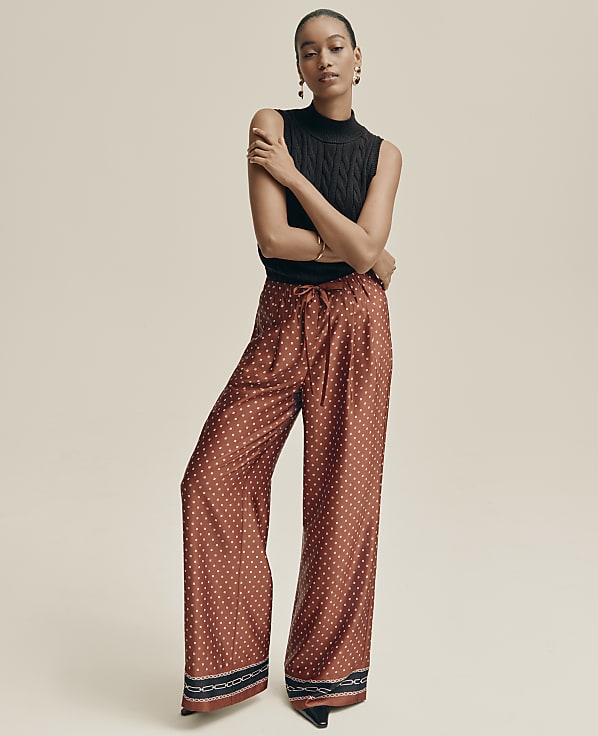 A woman in a black sleeveless turtleneck and patterned rust-colored wide-leg pants stands against a plain, light brown background.