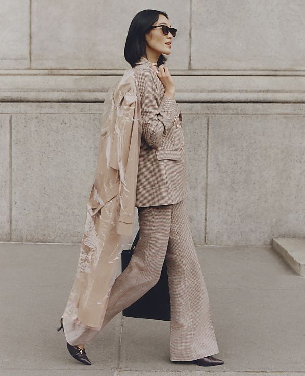 A woman in a tan plaid suit and sunglasses walks with a sheer trench coat draped over her shoulder in front of a stone building.