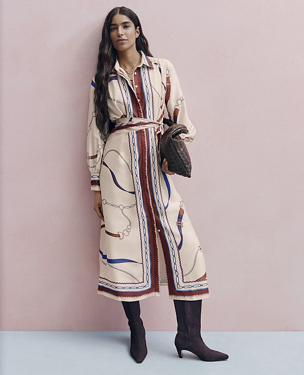 A woman in a patterned dress and boots stands against a pink wall, holding a dark purse.