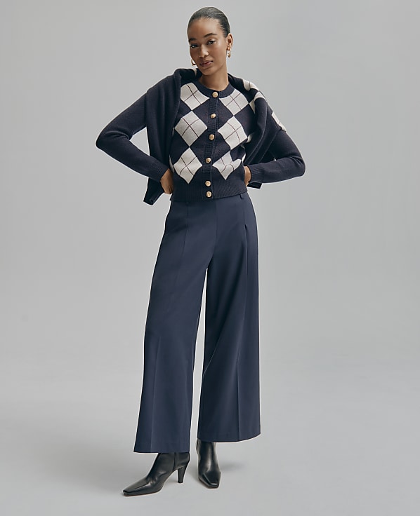 A woman in an argyle cardigan and wide-leg pants stands against a plain gray background.