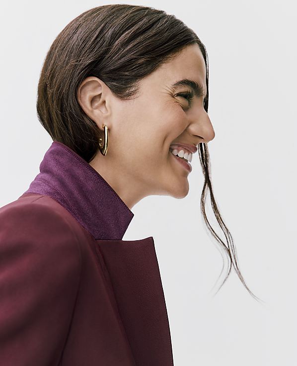 A woman with dark hair and a gold hoop earring smiles in profile, wearing a burgundy jacket with a purple collar against a plain white background.