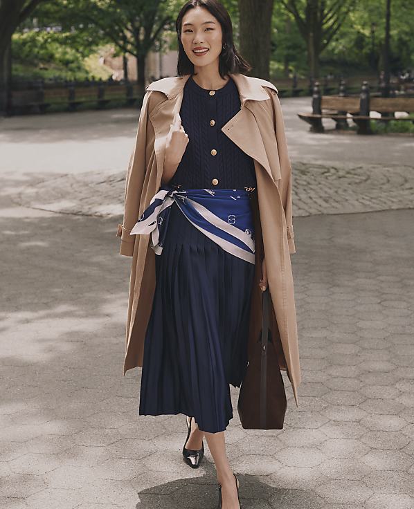 A woman in a trench coat and pleated skirt walks on a path in a park with benches and trees in the background.
