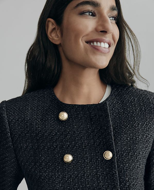 A smiling woman in a textured dark jacket with gold buttons is shown against a plain, light gray background.