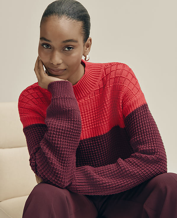 A woman in a red and burgundy sweater rests her chin on her hand against a light-colored background.