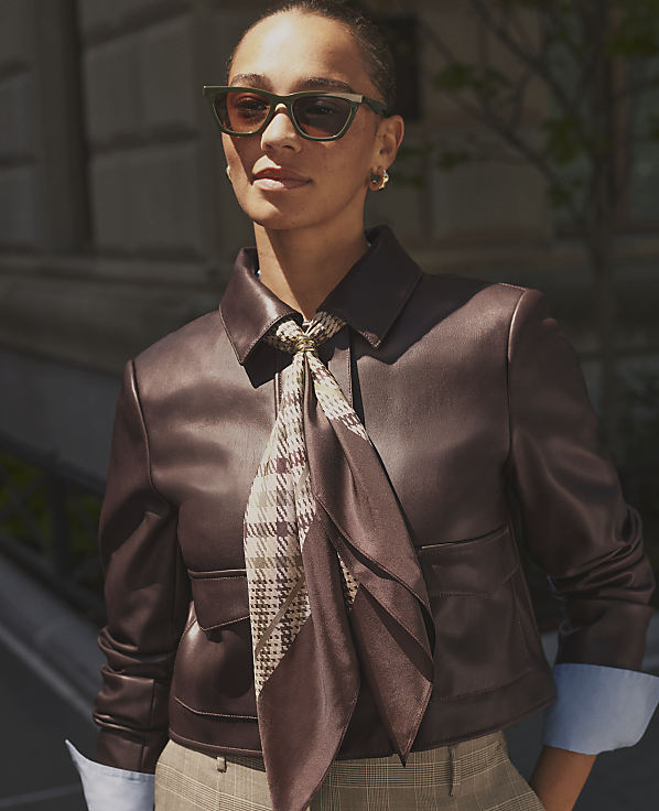 A woman in a brown leather jacket and plaid scarf stands in front of a blurred urban background.