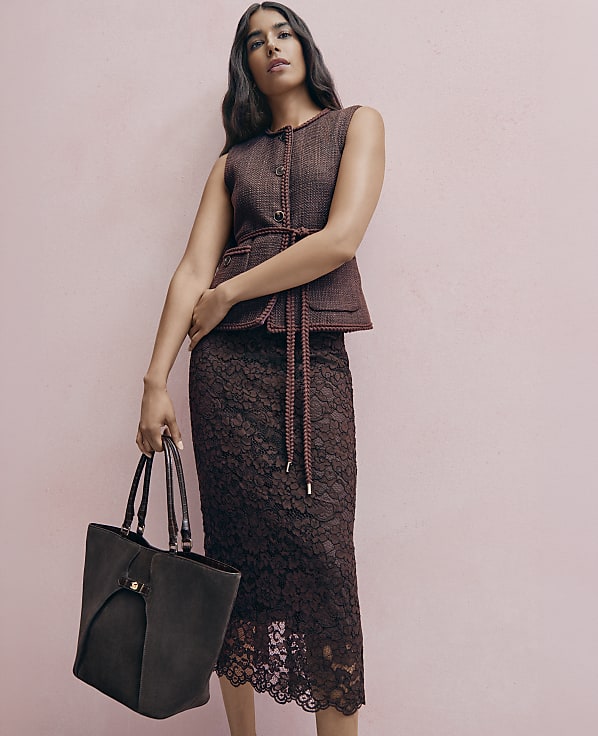 A woman in a brown tweed vest and lace skirt holds a dark tote bag against a soft pink background.