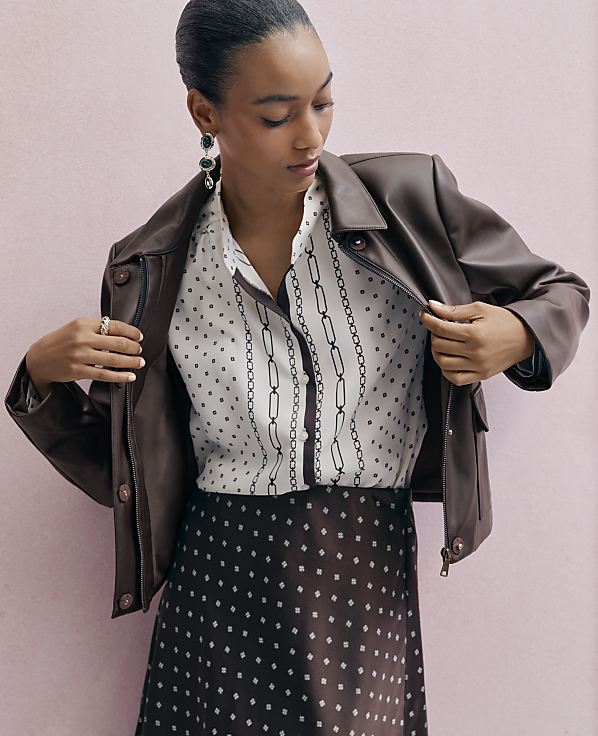 A woman in a patterned blouse and skirt, wearing a brown leather jacket, stands against a soft pink background.