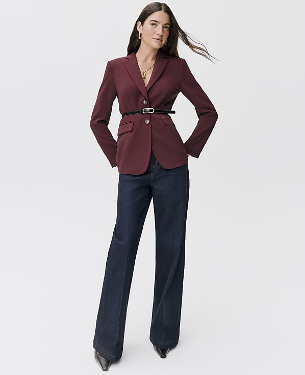 A woman in a burgundy blazer and dark wash jeans stands against a plain white background.