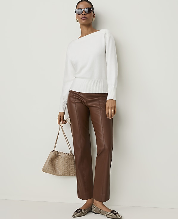A woman in a white sweater and brown leather pants stands against a plain white background, holding a beige handbag and wearing checkered flats.