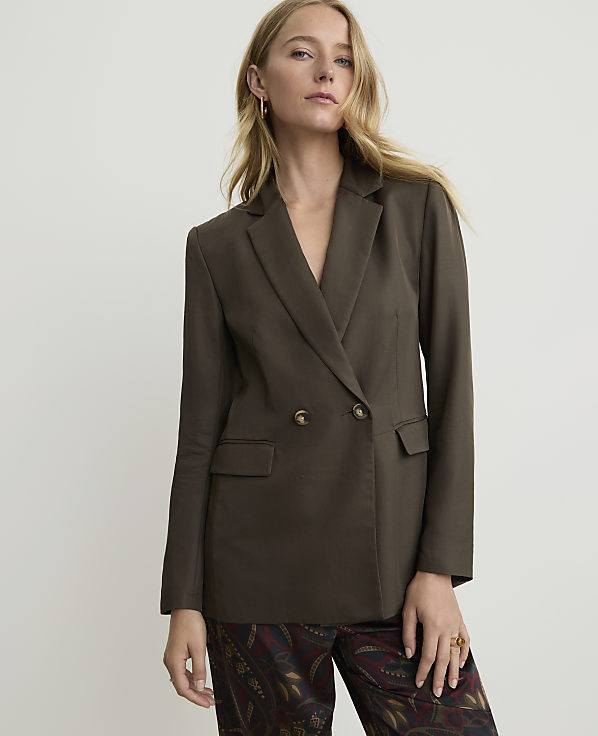 A woman in a dark brown double-breasted blazer and patterned pants stands against a plain white background.