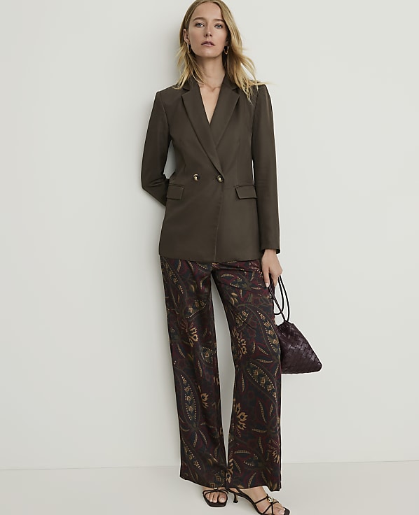 A woman in a brown blazer and patterned pants stands against a plain white wall, holding a small patterned bag.