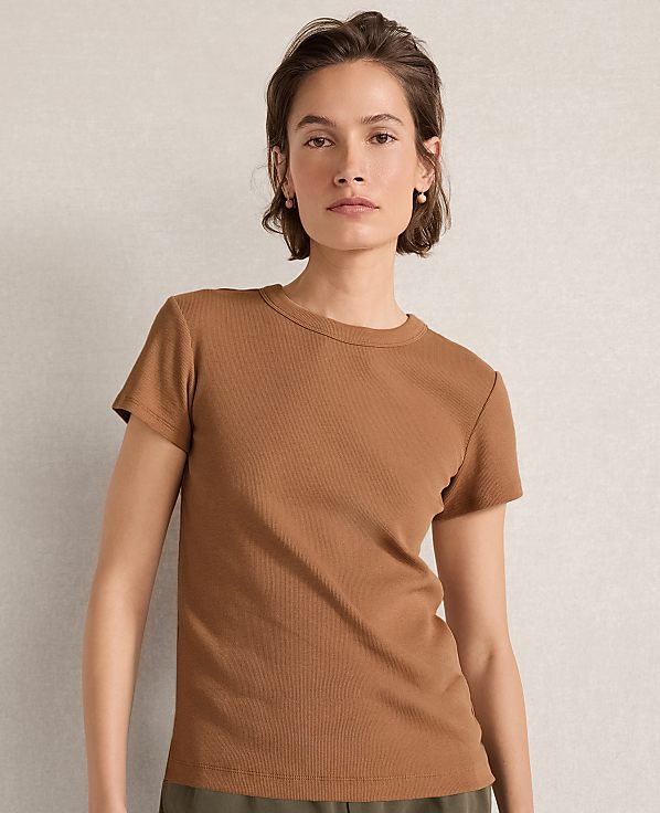 A woman in a brown ribbed t-shirt stands against a textured, neutral background.