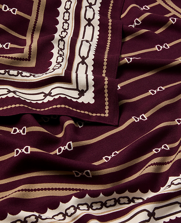 A folded burgundy scarf with a repeating equestrian bit pattern in beige and white is laid out, with a decorative border of chains and scallops visible on the left.