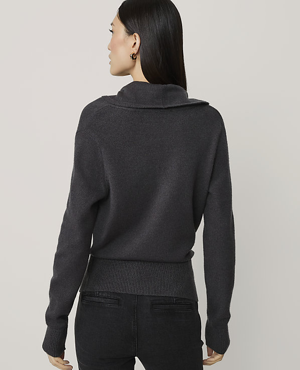 A woman in a dark grey sweater and black pants stands against a plain, light grey background.
