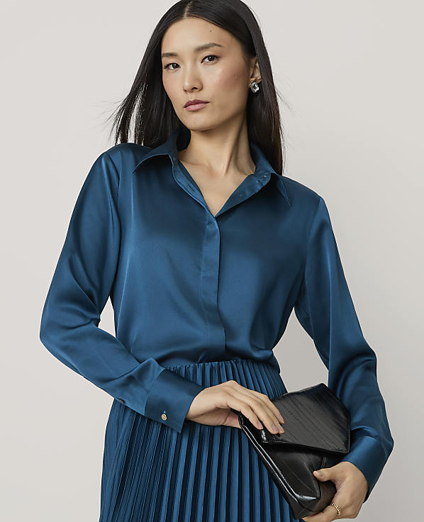A woman in a teal satin blouse and pleated skirt holds a black clutch against a plain background.