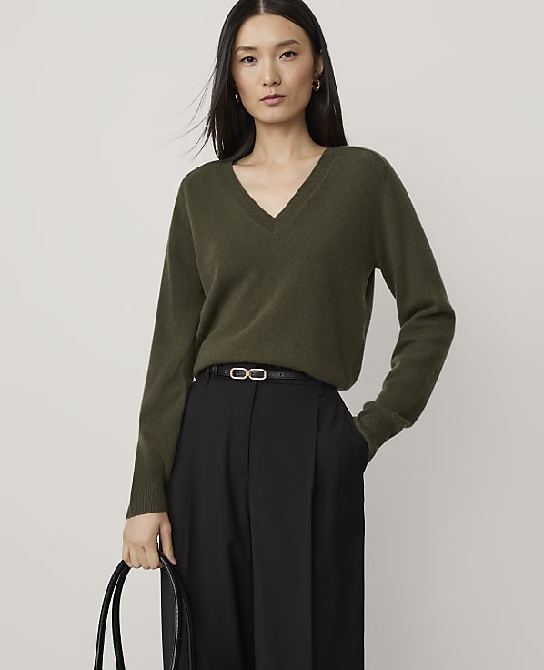 A woman in a green v-neck sweater and black pants stands against a plain, light background.
