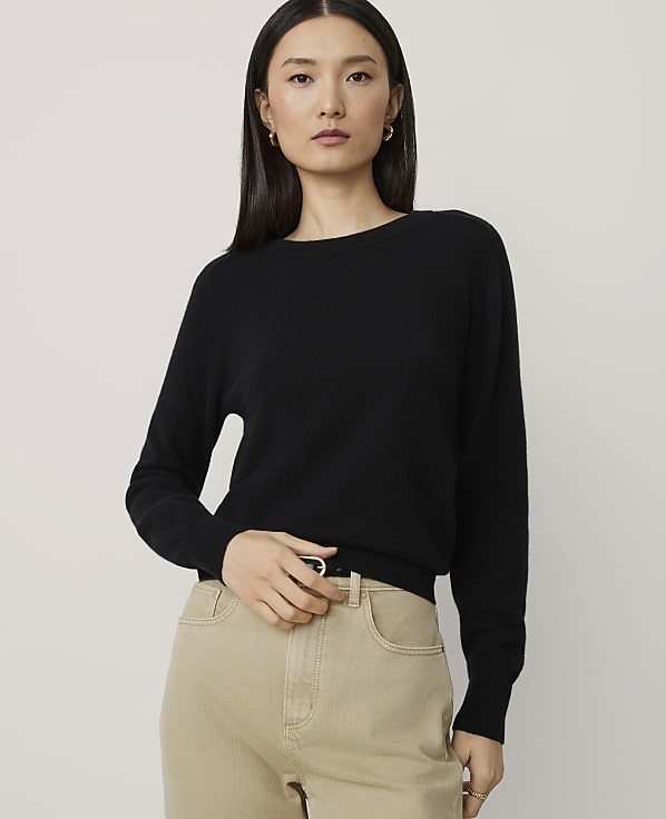 A woman in a black sweater and tan pants stands against a plain, light gray background.