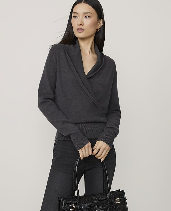 A woman in a dark grey wrap sweater and black jeans holds a black textured tote bag in front of a plain grey background.