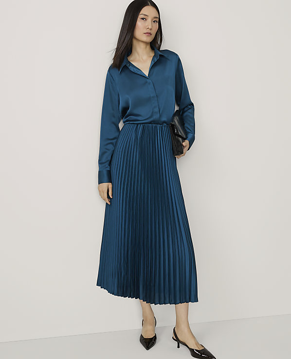 A woman in a teal satin blouse and pleated skirt stands against a plain white background, holding a black clutch.