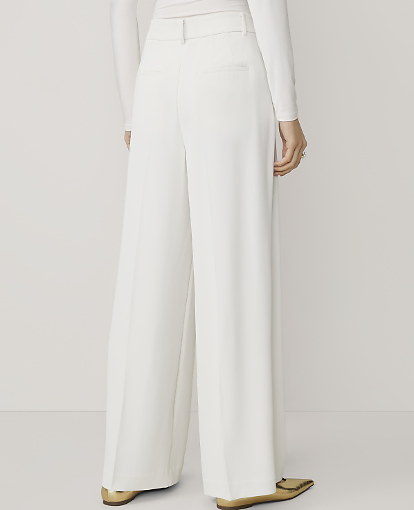 A person wearing white wide-leg pants and gold pointed-toe flats stands against a plain white background.