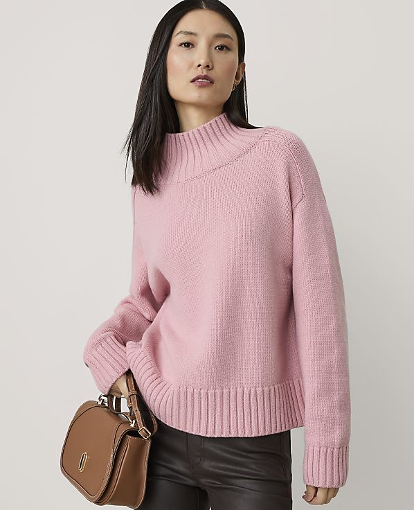 A woman in a pink sweater and dark pants holds a brown purse in front of a plain background.