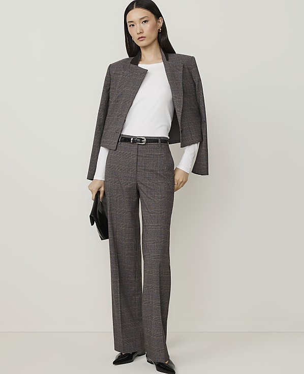 A woman in a grey plaid suit and black heels stands holding a dark woven clutch against a plain white background.