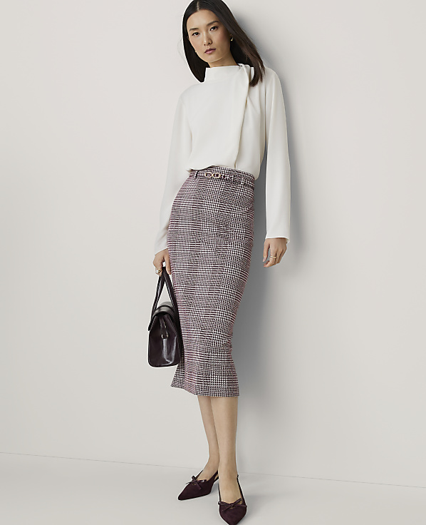 A woman in a white blouse and plaid skirt stands against a plain white background, holding a dark handbag.