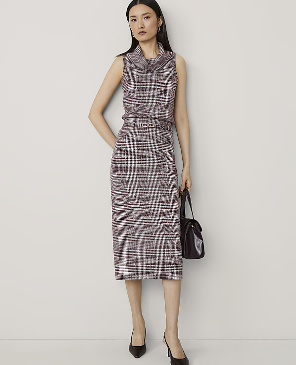 A woman in a plaid sleeveless turtleneck and skirt stands against a plain background, holding a dark handbag.